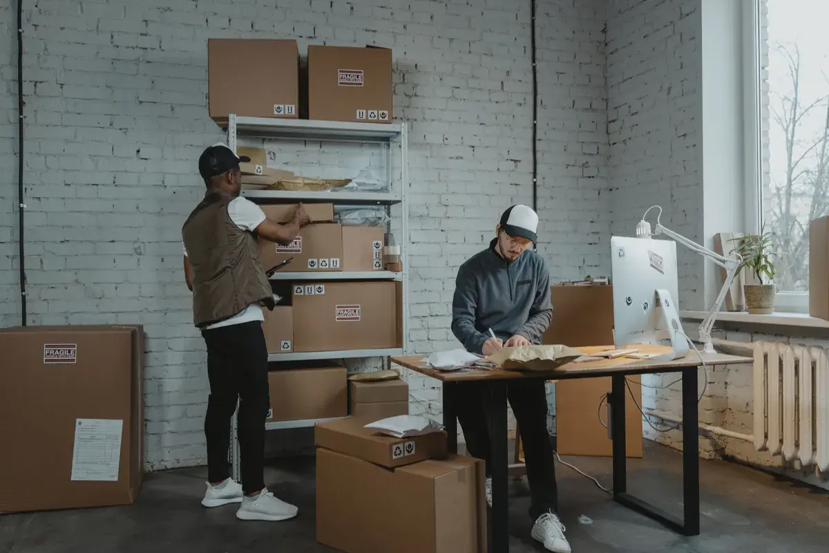 Two workers in a small shipping room, one writing on a form at a desk while the other shelves packages.