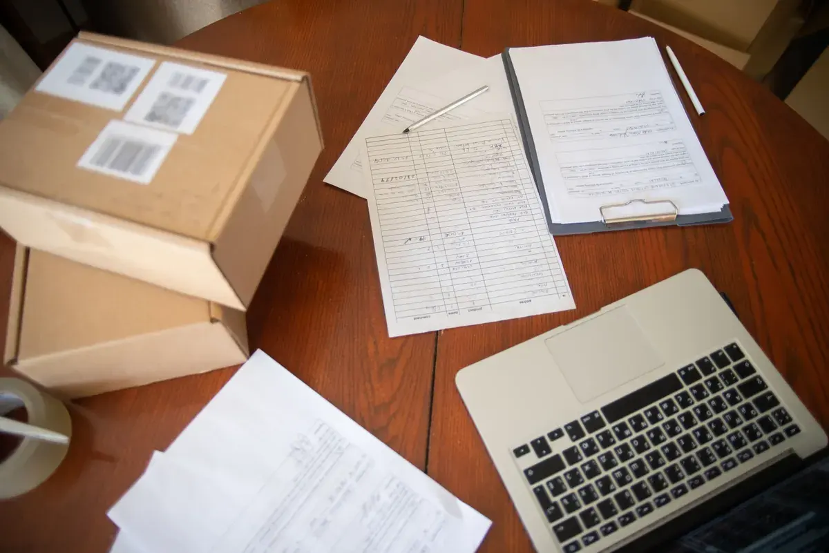 A wooden table with labeled parcels, handwritten order forms on a clipboard, and an open laptop.