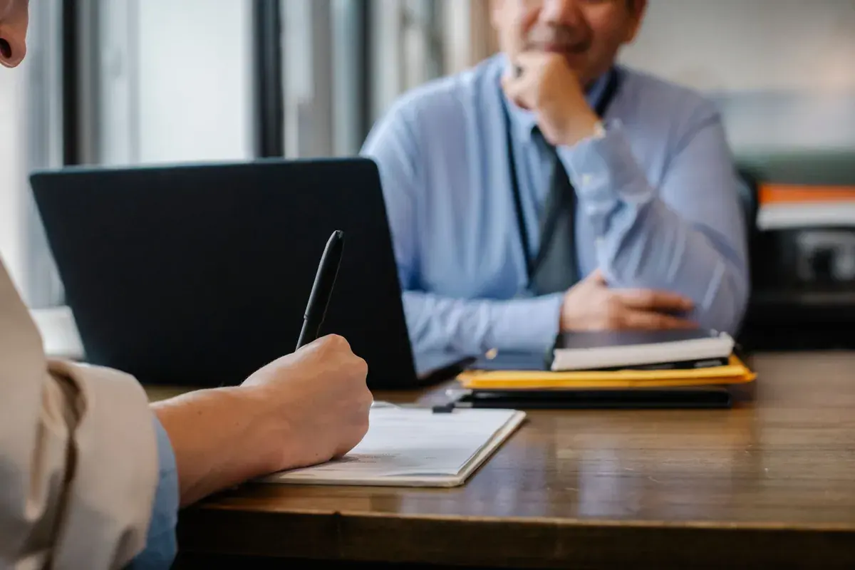 A person takes notes on a clipboard during an interview while another sits listening across the table.