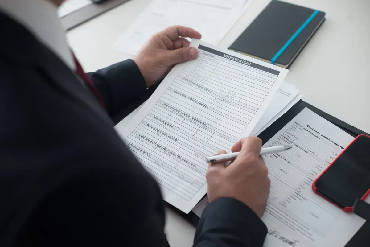 A person in a suit holds a printed application form over a desk, pen in hand, ready to complete it.